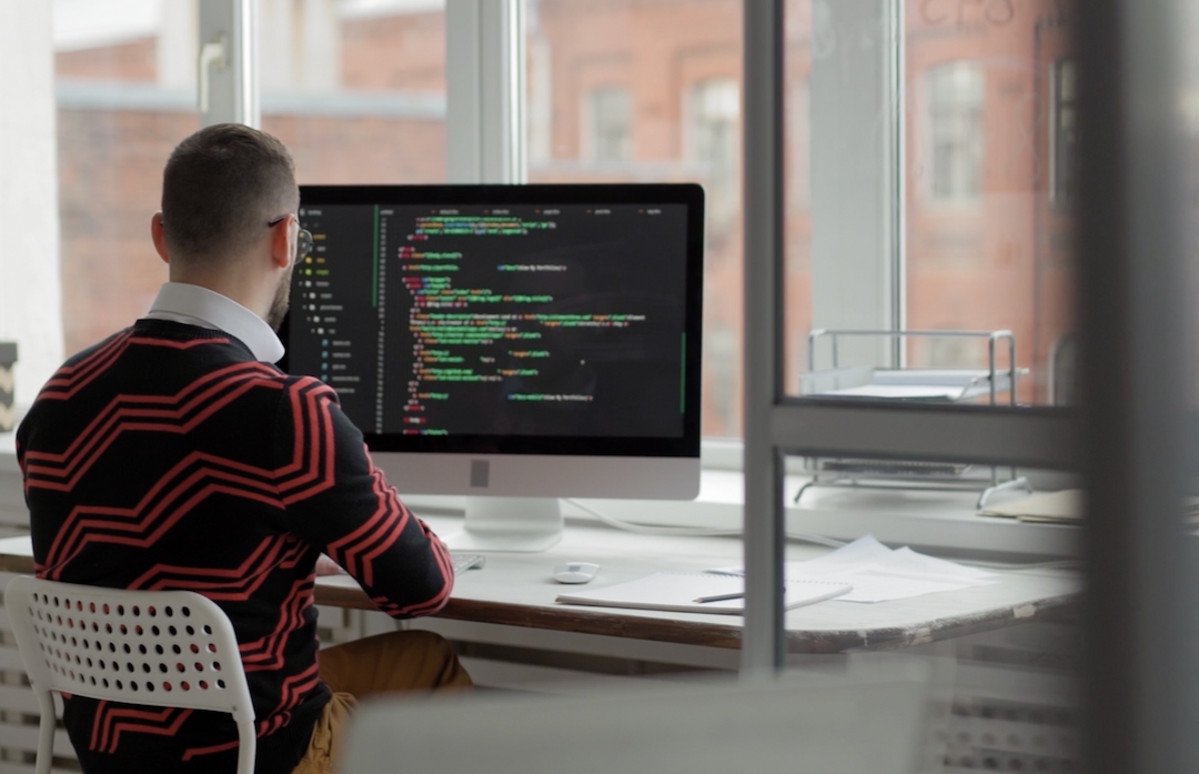 a view of a Target engineer dressed in a black sweater with red zig zag patterns looking at his screen with lines of code on it, in front of a window looking out on a city block of brick buildings