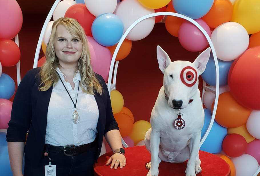 Target Senior Engineer Kaylee Edwards, with long blonde hair, smiling and wearing a white button down shirt and black cardigan. She is standing next to Target's mascot Bullseye, a white bull terrier with red Target logo around his eye. They appear in front of a background of colorful balloons.