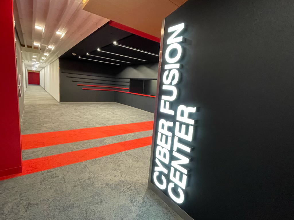 photo of Target's Cyber Fusion Center entrance, with illuminated white letters mounted vertically to a black painted wall, and red striped carpet with a long hallway in the background